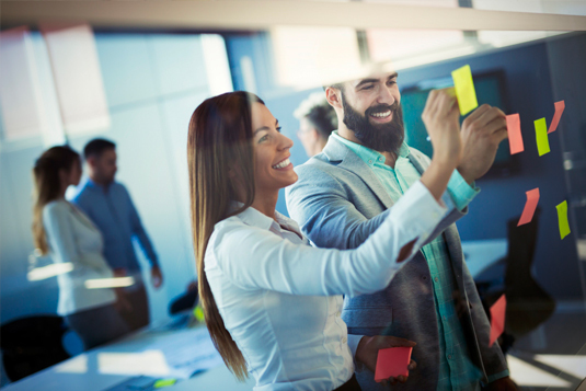 Smiling colleagues placing sticky notes on a glass wall during a team brainstorm