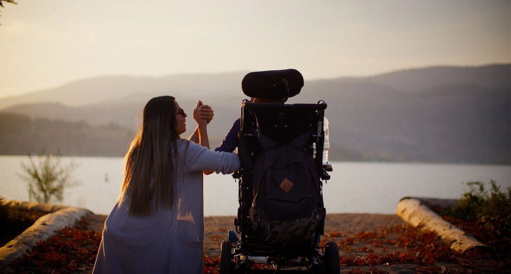 A woman shares a quiet moment with a person in a power wheelchair beside a lake at sunset