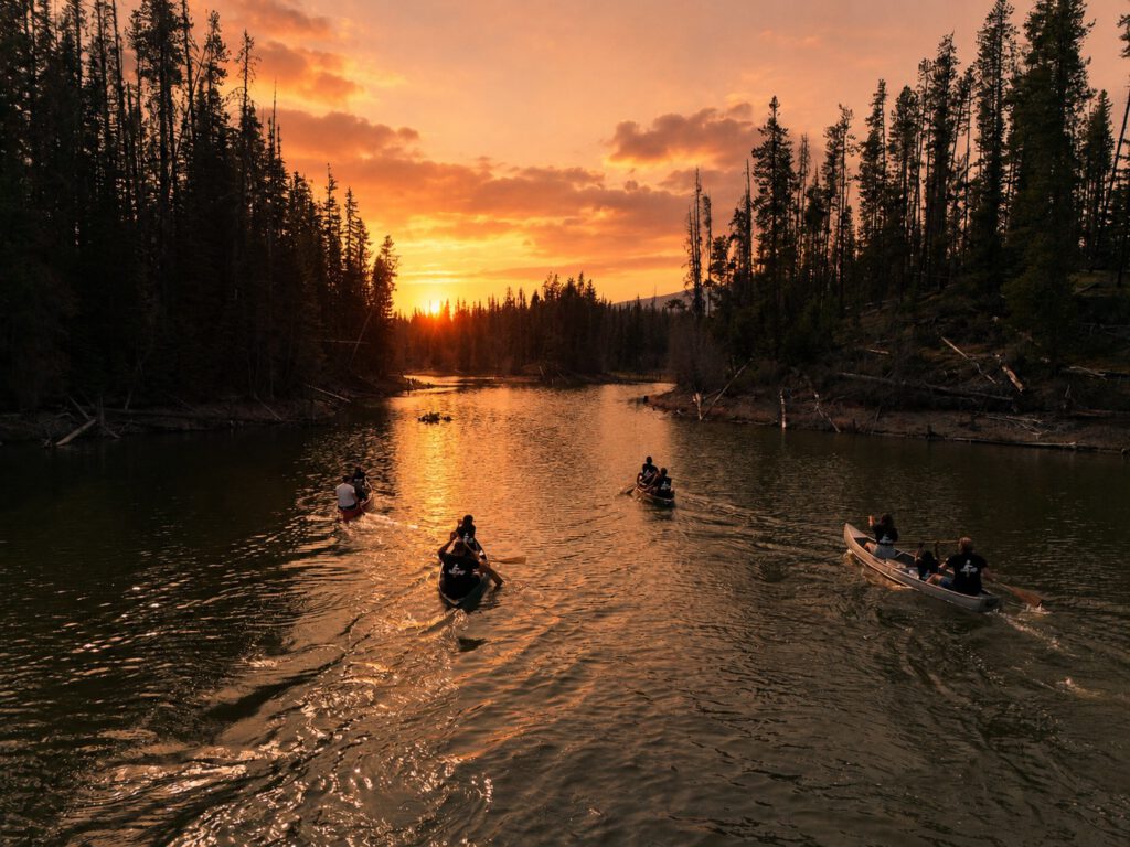 Canoeists paddle down a river toward a glowing orange sunset
